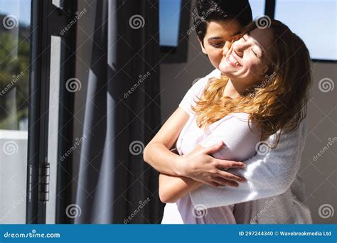 Happy Biracial Lesbian Couple Embracing In Sun By Window At Home Copy