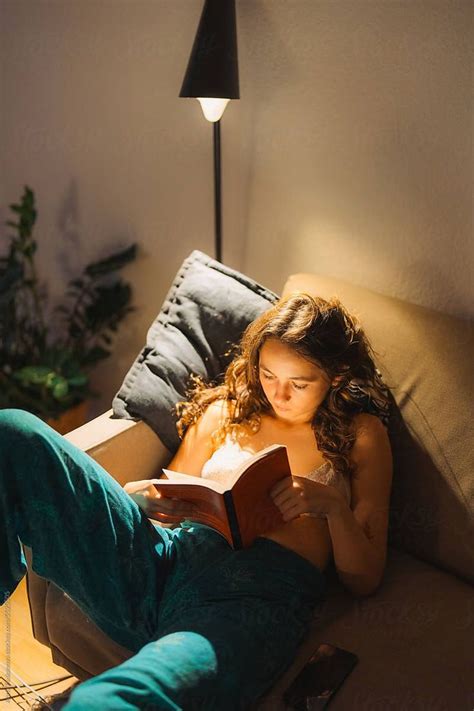 Woman Laying On The Couch Reading Book In The Evening By Stocksy