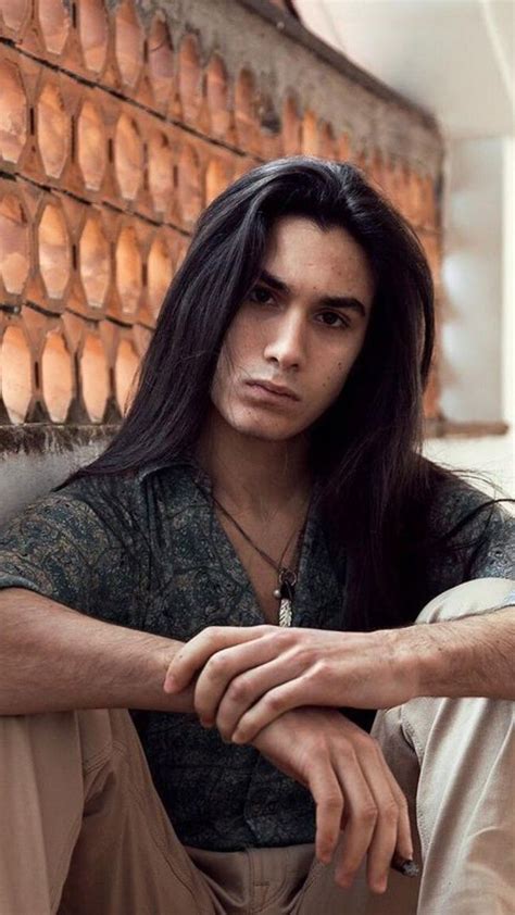 A Man With Long Black Hair Sitting In Front Of A Wall