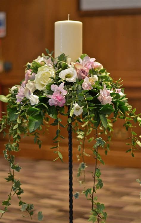 A White Candle Is Sitting On Top Of A Black Stand With Flowers And Greenery