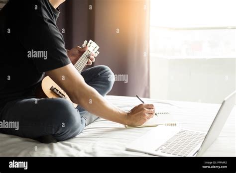 Happy Young Asian Man Playing Ukulele Sitting On Bed In Bedroom Stock