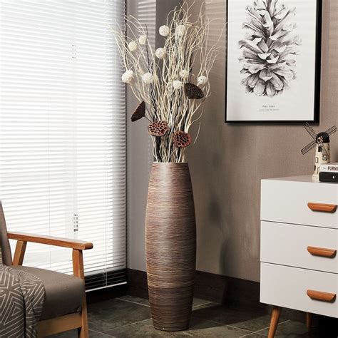 A Tall Vase With Flowers In It Next To A Chair And Pictures On The Wall