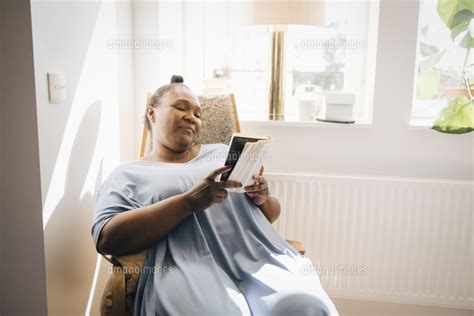 Smiling Woman Reading Book While Sitting On Chair In Living Room