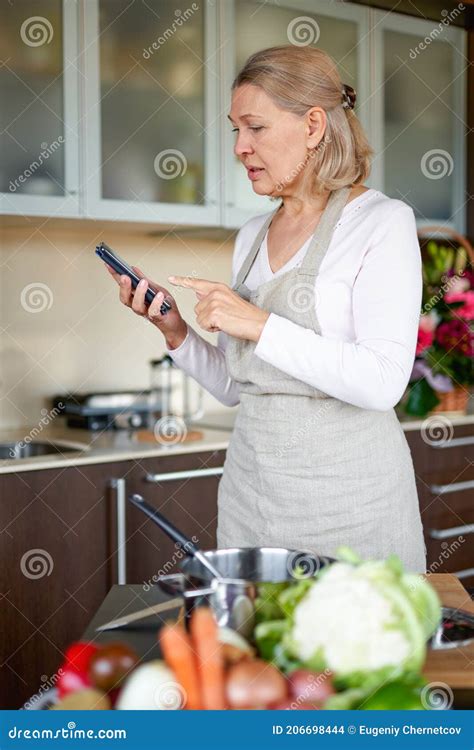 Mature Woman In Kitchen Preparing Food And Holding Smart Phone Stock
