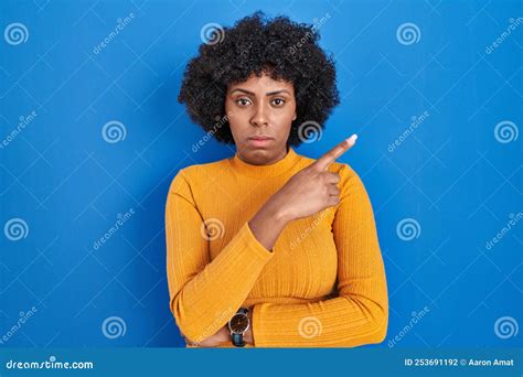 Black Woman With Curly Hair Standing Over Blue Background Pointing With Hand Finger To The Side