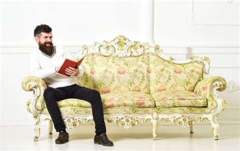 Man With Beard And Mustache Sits On Baroque Style Sofa Holds Book