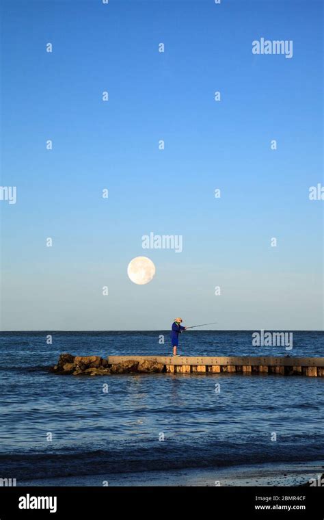 Fisherman On The Jetty As The Full Moon Sets Over The Ocean In Miami