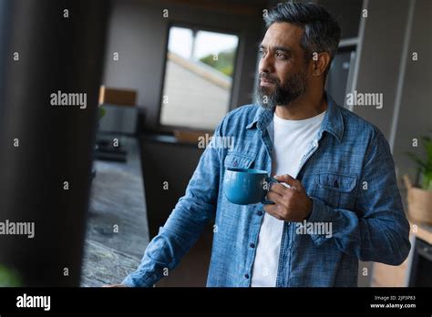 Happy Biracial Man Holding Mug And Looking Through Window In Kitchen