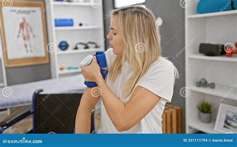 A Blonde Woman Exercises Her Arm With A Dumbbell In A Clinic S Rehab Room Surrounded By Medical